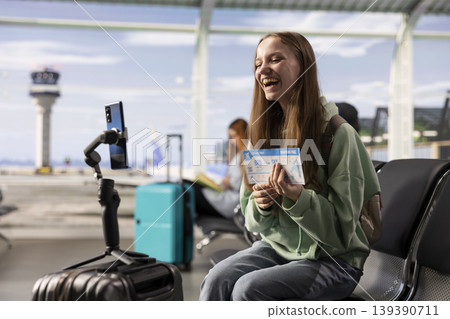 Girl content creator using phone on tripod to record vlog in airport lounge, capturing travel journey before boarding. Busy terminal illustrates social media presence and mobile filming. Girl content creator using phone on tripod to record vlog in airport lounge, capturing travel journey before boarding. Busy terminal illustrates social media presence and mobile filming. 139390711