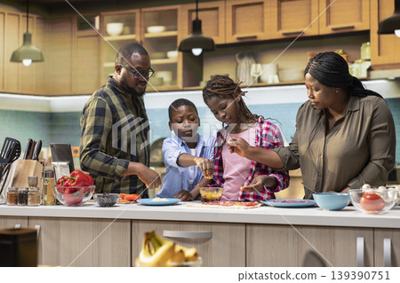 Happy black parents teaching children to cook and cut vegetables, enjoy making homemade pizza from old family recipe. Mom and dad explaining steps for efficient cooking in the kitchen. 139390751