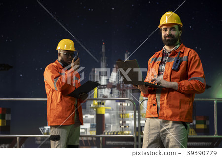 Portrait of smiling offshore platform engineer using laptop to oversee drilling during night shift. Happy drilling rig professional looking over sensor data to read production metrics on notebook Portrait of smiling offshore platform engineer using laptop to oversee drilling during night shift. Happy drilling rig professional looking over sensor data to read production metrics on notebook 139390779