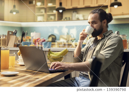 Adult man, morning coffee, working from home, kitchen environment, laptop for online work. Remote worker lifestyle, wireless internet and modern technology, multitasking, digital nomad. 139390798