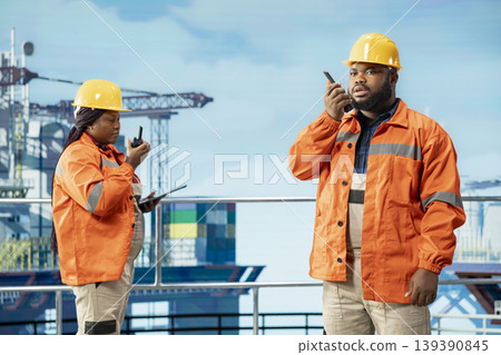 Drilling rig engineering team uses two way radio to communicate with logistics crew. Offshore platform teamworking colleagues speak in walkie talkie providing audio clarity in noisy environment 139390845