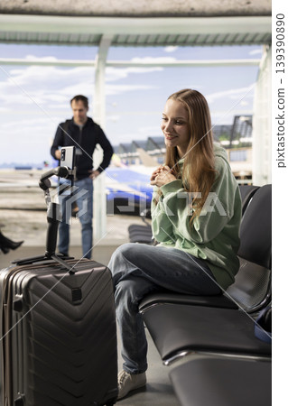Young tourist, woman filming travel vlog, in the airport lounge, smiling and talking to audience through mobile phone. Content creator highlights streaming and the flexibility of working online. Young tourist, woman filming travel vlog, in the airport lounge, smiling and talking to audience through mobile phone. Content creator highlights streaming and the flexibility of working online. 139390890