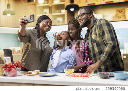 Smiling mom takes selfies on smartphone with family while cooking delicious homemade pizza together in kitchen, children enjoying laughter and the warmth of bonding with their parents. 139390960