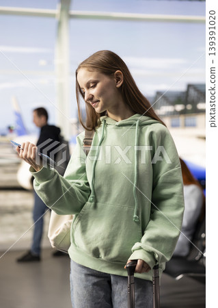 Gen z girl traveler in airport waiting area verifies information on plane ticket, busy terminal activity with baggage and passengers. Tourism, economy travel and real-life airport experiences. 139391020