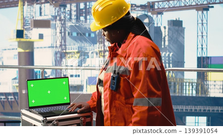 Green screen laptop used by drilling rig worker holding handheld radio device to monitor operation. Woman on offshore platform deck using walkie talkie and mockup notebook, calibrating gear 139391054