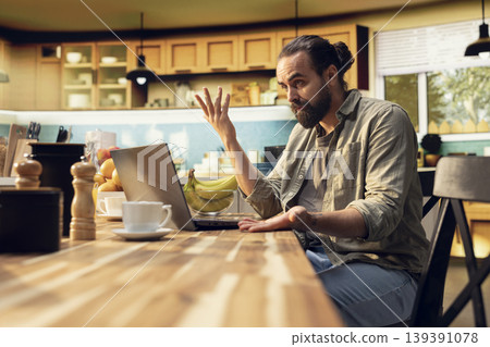 Uncertain freelancer being confused about ambiguous tasks at home, remote work on laptop in the kitchen. Disoriented disorganized man unsure about telecommuting job, frowning. 139391078