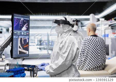 Female specialist in protective gear checking DNA molecules of infected patient on monitor in lab, analyzing pathology and virology data, testing immunology theories and developing vaccines. Female specialist in protective gear checking DNA molecules of infected patient on monitor in lab, analyzing pathology and virology data, testing immunology theories and developing vaccines. 139391129