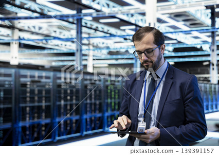 Data center system administrator using tablet to monitor performance, identifying bottlenecks. Server farm IT specialist using device app to implement optimizations improving electronics 139391157