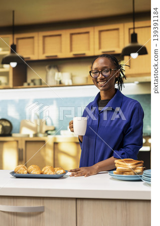 Portrait of jolly african american woman serving coffee in a mug before eating bread slices and pastry for breakfast meal. Enjoying calm morning surrounded by kitchenware and healthy food. Portrait of jolly african american woman serving coffee in a mug before eating bread slices and pastry for breakfast meal. Enjoying calm morning surrounded by kitchenware and healthy food. 139391184