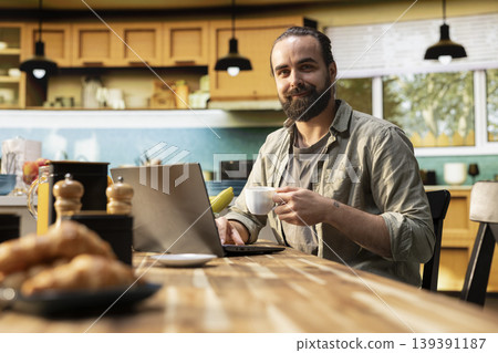 Portrait of teleworker drinking his morning coffee with laptop at kitchen table, routine with daily teleworking tasks for relaxed lifestyle and productivity. Working from home indoors. Portrait of teleworker drinking his morning coffee with laptop at kitchen table, routine with daily teleworking tasks for relaxed lifestyle and productivity. Working from home indoors. 139391187