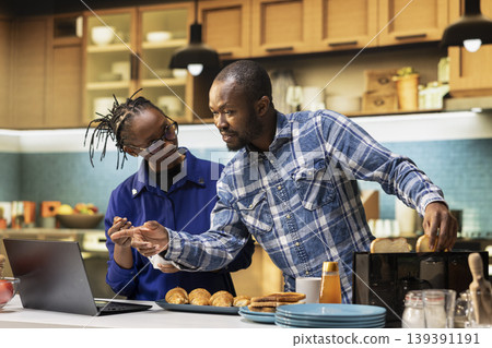 Jolly boyfriend and girlfriend taking bread out of the toaster for breakfast meal, standing at kitchen counter and laughing. Couple preparing delicious food in the morning meal with coffee cups. Jolly boyfriend and girlfriend taking bread out of the toaster for breakfast meal, standing at kitchen counter and laughing. Couple preparing delicious food in the morning meal with coffee cups. 139391191