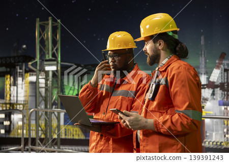 Offshore platform crew members using laptop to control subsea systems late at night. Teamworking colleagues monitoring anomaly detection using notebook on drilling derrick 139391243
