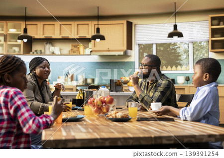 African American family sharing pastry and drinks for breakfast at home, parents serve scrambled eggs, toast and fruit. Cozy kitchen moment captures warmth, relaxation and joyful togetherness. 139391254