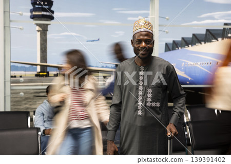 Portrait of smiling man in airport wearing traditional clothing and blurry crowd traveling by plane during holiday trip. Happy African person in ethnic attire and silhouettes of travelers walking Portrait of smiling man in airport wearing traditional clothing and blurry crowd traveling by plane during holiday trip. Happy African person in ethnic attire and silhouettes of travelers walking 139391402