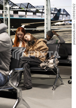 Woman in airport resting head on friend shoulder, feeling upset after delayed flight. Sad girl comforted by sister after losing flight due to airline overbooking, waiting for another plane 139391422