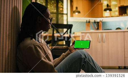 Mockup chroma key on mobile phone held by young black woman in cozy home, sitting on the floor in comfort under soft light. Female adult in a peaceful everyday lifestyle setting. Mockup chroma key on mobile phone held by young black woman in cozy home, sitting on the floor in comfort under soft light. Female adult in a peaceful everyday lifestyle setting. 139391526
