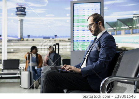 Entrepreneur traveling, international business trip, sitting in airport lounge, with baggage and laptop open. Businessman studies documents and plans while passengers move around, departure area. Entrepreneur traveling, international business trip, sitting in airport lounge, with baggage and laptop open. Businessman studies documents and plans while passengers move around, departure area. 139391546