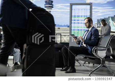 Entrepreneur in corporate attire at the airport waiting area near gate, holding boarding pass and passport and checking flight information on screen. International business travel and mobility. Entrepreneur in corporate attire at the airport waiting area near gate, holding boarding pass and passport and checking flight information on screen. International business travel and mobility. 139391547