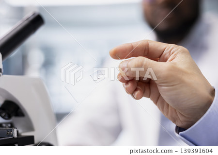 Close up of molecular biologist examining microscope glass slide in laboratory to view cells. Research facility professional checks sample slide before imaging process using optical device 139391604
