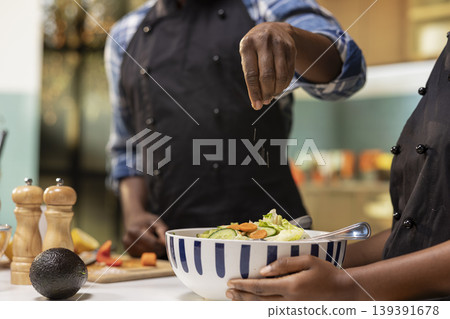 Close up of african american couple preparing food together in their kitchen, adding a pinch of salt to a fresh summer salad bowl. Man and woman enjoying a healthy and organic culinary activity. Close up of african american couple preparing food together in their kitchen, adding a pinch of salt to a fresh summer salad bowl. Man and woman enjoying a healthy and organic culinary activity. 139391678