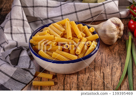 Uncooked Rigatoni Pasta in Ceramic Bowl. Kitchen Cloth, Garlic, Red Pepper on Rustic Wooden Table 139395469