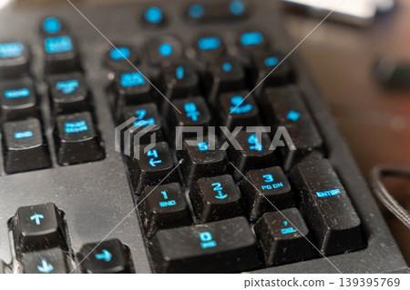 Close-up view of a black computer keyboard with blue backlit keys, showing dust accumulation and various function keys, placed on a wooden desk surface Close-up view of a black computer keyboard with blue backlit keys, showing dust accumulation and various function keys, placed on a wooden desk surface 139395769