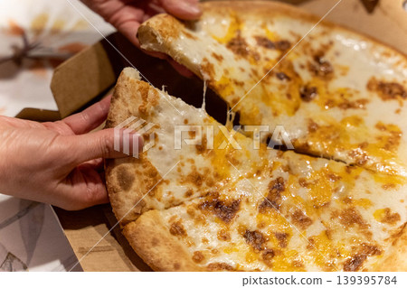 Hands pulling apart a cheesy pizza slice from a pizza box, with melted cheese stretching and a blurred tablecloth in the background Hands pulling apart a cheesy pizza slice from a pizza box, with melted cheese stretching and a blurred tablecloth in the background 139395784