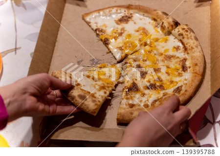 Hand reaching for a slice of cheese pizza from an open cardboard box on a table with a floral tablecloth and a partially eaten pizza visible Hand reaching for a slice of cheese pizza from an open cardboard box on a table with a floral tablecloth and a partially eaten pizza visible 139395788