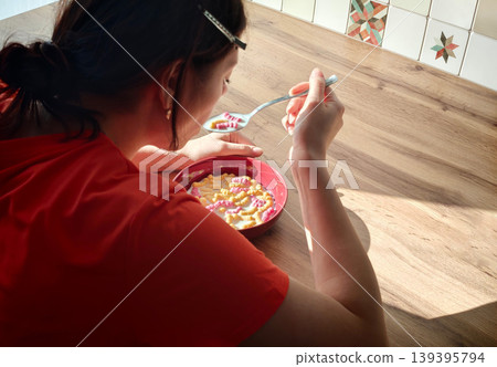 Woman with dark hair in red shirt enjoys breakfast cereal from a red bowl while seated at a wooden table with patterned tiles in the background 139395794