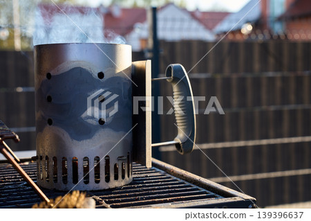 Metal charcoal chimney starter releases smoke on an outdoor grill grate 139396637
