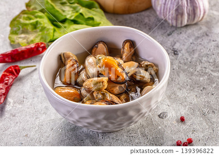 Marinated mussels in a white bowl with chili peppers, garlic, and fresh lettuce on a concrete background. 139396822