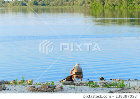 Man sitting on a rocky beach fishing into a calm lake on a sunny day with trees in the background 139397058