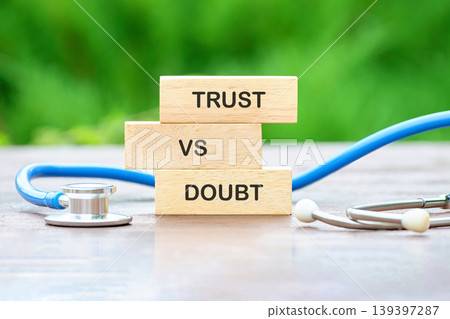 Three wooden blocks displaying TRUST VS DOUBT are stacked vertically. A blue medical stethoscope rests beside them on a dark wooden table, set against a blurred green background 139397287