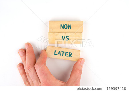 A hand holds a wooden block labeled Later, positioned beneath two stacked blocks that read Now and VS A hand holds a wooden block labeled Later, positioned beneath two stacked blocks that read Now and VS 139397418
