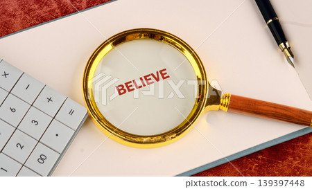 A magnifying glass highlights the word BELIEVE on white paper. A calculator and fountain pen rest nearby on a textured surface A magnifying glass highlights the word BELIEVE on white paper. A calculator and fountain pen rest nearby on a textured surface 139397448