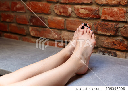 Woman bare legs and feet with black toenail polish on a concrete floor against a brick wall Woman bare legs and feet with black toenail polish on a concrete floor against a brick wall 139397608