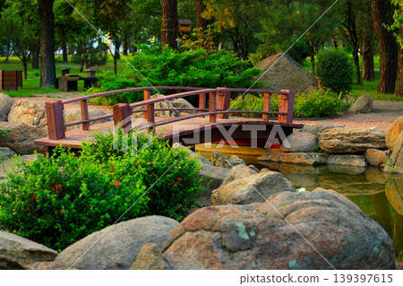 Japanese garden with a wooden bridge over a pond surrounded by large rocks and lush greenery 139397615