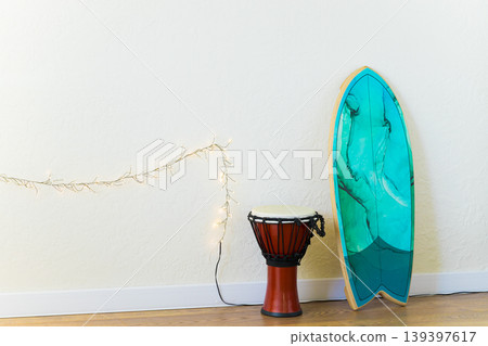 djembe drum and a turquoise surfboard on a wooden floor next to a white wall with string lights 139397617