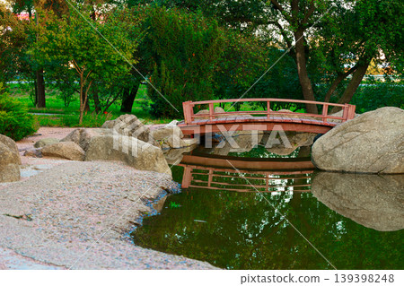 Asia Japanese style garden scene featuring a red wooden bridge over a pond surrounded by large rocks and lush greenery Asia Japanese style garden scene featuring a red wooden bridge over a pond surrounded by large rocks and lush greenery 139398248