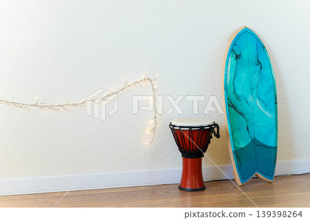 A djembe drum and a blue surfboard leaning against a white wall with string lights A djembe drum and a blue surfboard leaning against a white wall with string lights 139398264