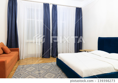 Modern bedroom with orange sofa, blue bed, and patterned rug near window Modern bedroom with orange sofa, blue bed, and patterned rug near window 139398273