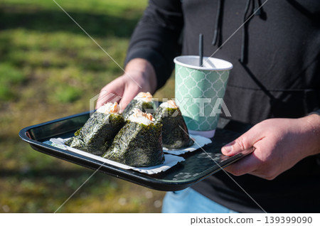 Closeup of compact lunch with beverage. Hand holding small tray featuring onigiri and cup detail Closeup of compact lunch with beverage. Hand holding small tray featuring onigiri and cup detail 139399090