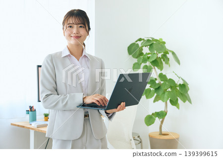 A young female business person operating a laptop computer; an office worker standing and working in the office. A young female business person operating a laptop computer; an office worker standing and working in the office. 139399615