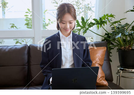 一位年輕、微笑的職業女性穿著套裝,正在遠端辦公,她坐在沙發上使用筆記型電腦。 一位年輕、微笑的職業女性穿著套裝,正在遠端辦公,她坐在沙發上使用筆記型電腦。 139399695