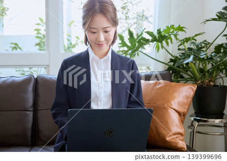 一位身著職業套裝的年輕日本女商人坐在沙發上用筆記型電腦工作——這是遠距辦公的典型畫面。 139399696