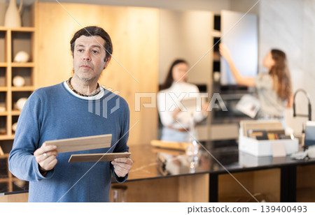 Male buyer with samples of material chooses kitchen set in furniture store Male buyer with samples of material chooses kitchen set in furniture store 139400493