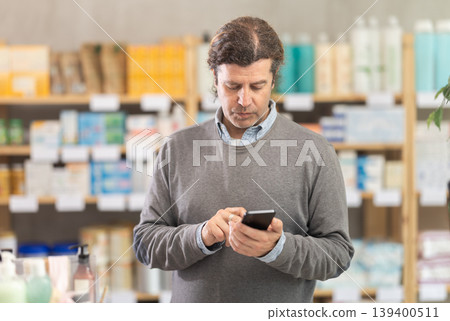 Male shopper searches for necessary goods on the shelves of a supermarket, checking list on her smartphone 139400511