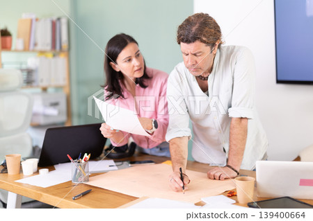 Middle-aged colleagues discussing construction plan leaning over office table Middle-aged colleagues discussing construction plan leaning over office table 139400664