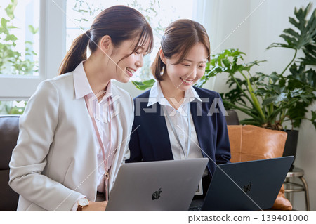 Two young female business professionals are seen smiling and having a meeting while looking at their laptops – a collaborative scene in an office. Two young female business professionals are seen smiling and having a meeting while looking at their laptops – a collaborative scene in an office. 139401000