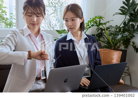 兩位年輕的職業婦女在辦公室休息室裡，打開筆記型電腦，查看手錶上的時間。 139401002